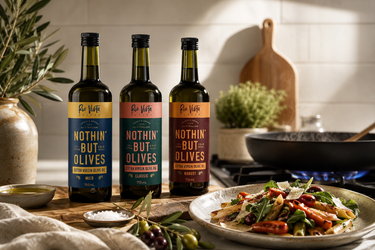 Three bottles of Rio Vista 'Nothin' But Olives' extra virgin olive oil in different flavours displayed on a rustic kitchen counter next to a prepared pasta dish, with a stove and tiled background.