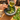 A close-up of hands preparing a green sauce in a stone mortar and pestle, pouring olive oil over chopped herbs and cheese on a wooden board, with fresh herbs and grated cheese nearby.