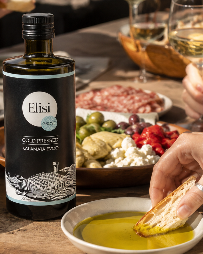 Bottle of Elisi Kalamata EVOO with a person dipping bread into olive oil on a table with food.